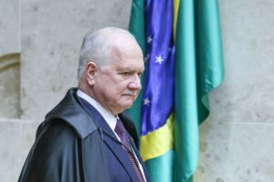 Um homem de toga caminha em frente a uma bandeira verde e amarela com estrelas e faixa azul