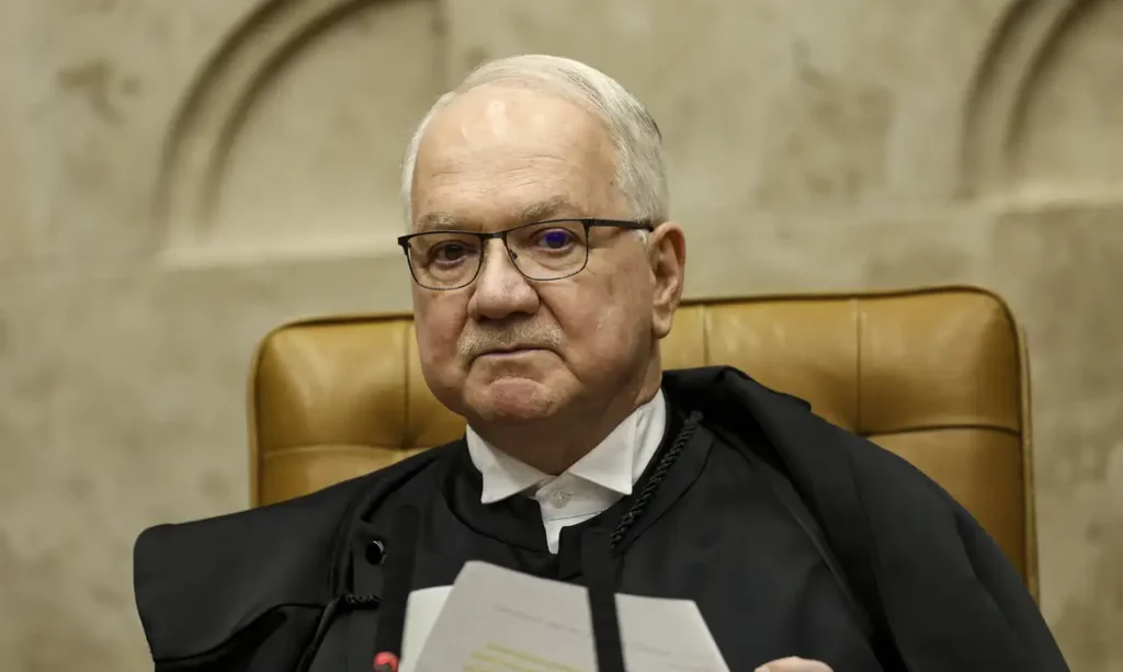 Um homem de toga e óculos sentado em cadeira de couro segura documentos em ambiente formal