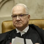 Um homem de toga e óculos sentado em cadeira de couro segura documentos em ambiente formal