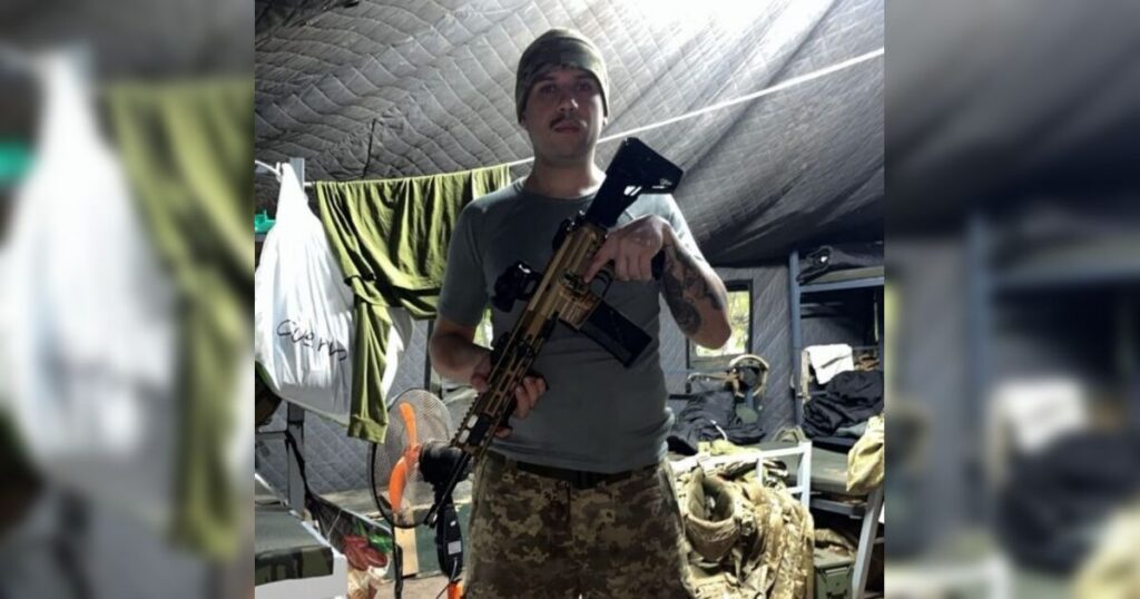 Um homem segura uma arma dentro de um abrigo com camas e roupas penduradas ao fundo