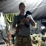 Um homem segura uma arma dentro de um abrigo com camas e roupas penduradas ao fundo