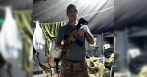 Um homem segura uma arma dentro de um abrigo com camas e roupas penduradas ao fundo