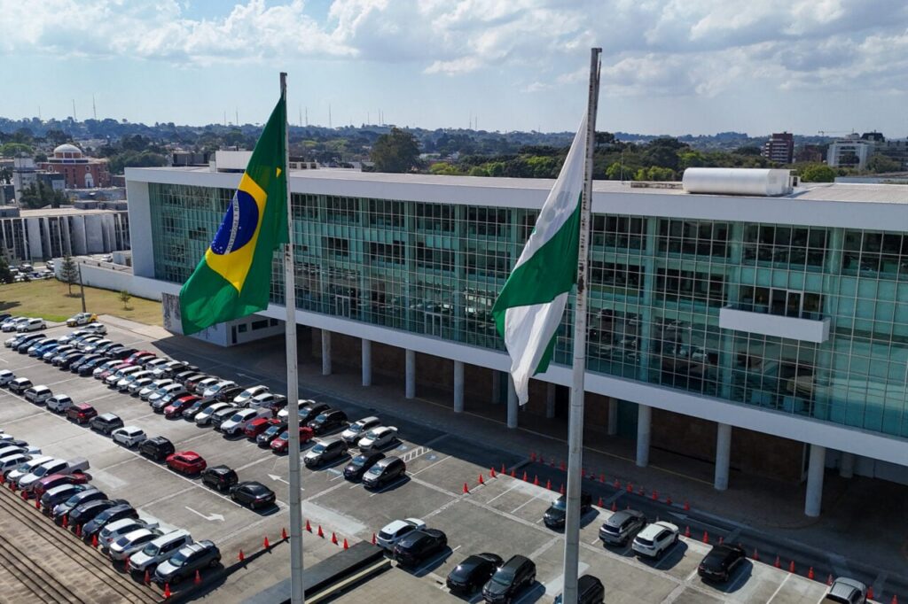 Prédio moderno com fachada de vidro e bandeiras hasteadas, cercado por estacionamento com diversos veículos