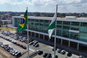 Prédio moderno com fachada de vidro e bandeiras hasteadas, cercado por estacionamento com diversos veículos