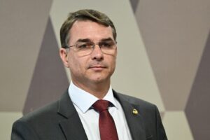 Um homem de terno e gravata vermelha com óculos olha para frente em frente a parede com padrão geométrico