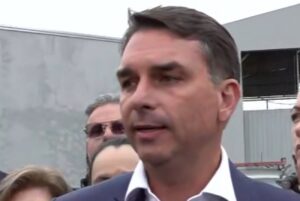 Um homem de terno e camisa branca fala em frente a um prédio com outras pessoas ao fundo
