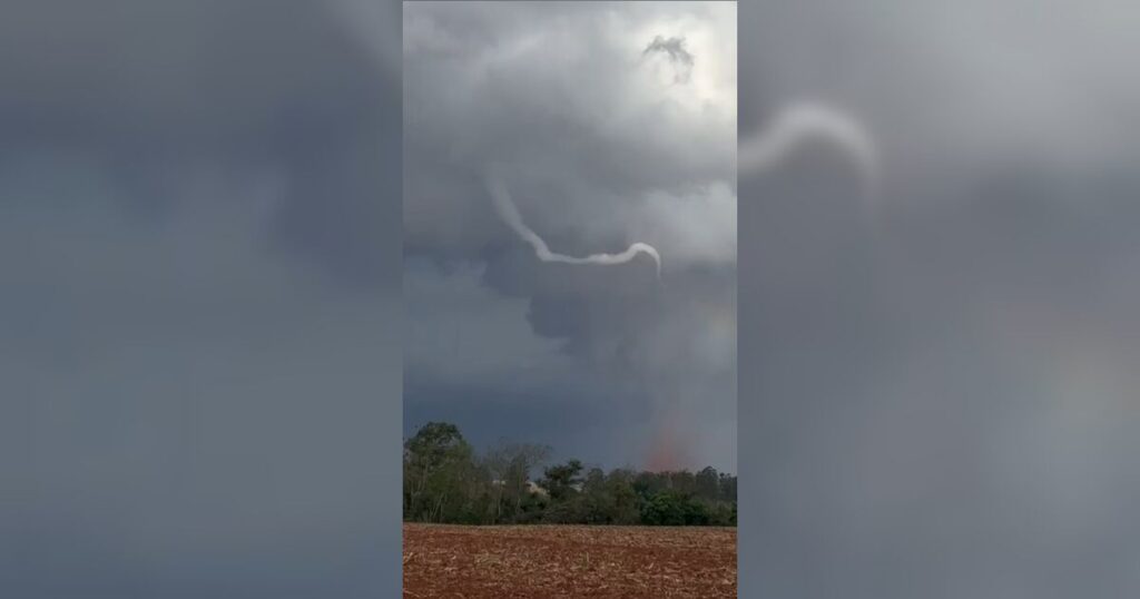 Fundo de campo com terra arada e vegetação, sob nuvens escuras e um funil de tornado se formando no céu