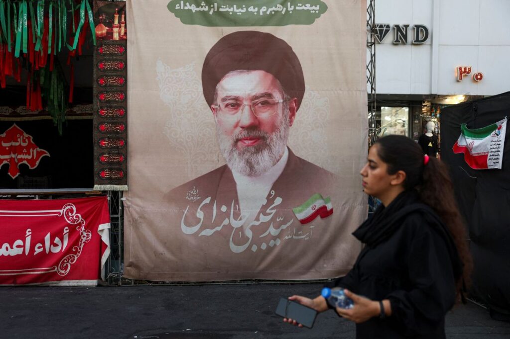 Uma mulher caminha em frente a um grande cartaz com retrato de um homem de turbante e barba, em meio a bandeiras e faixas com inscrições em persa