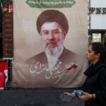 Uma mulher caminha em frente a um grande cartaz com retrato de um homem de turbante e barba, em meio a bandeiras e faixas com inscrições em persa