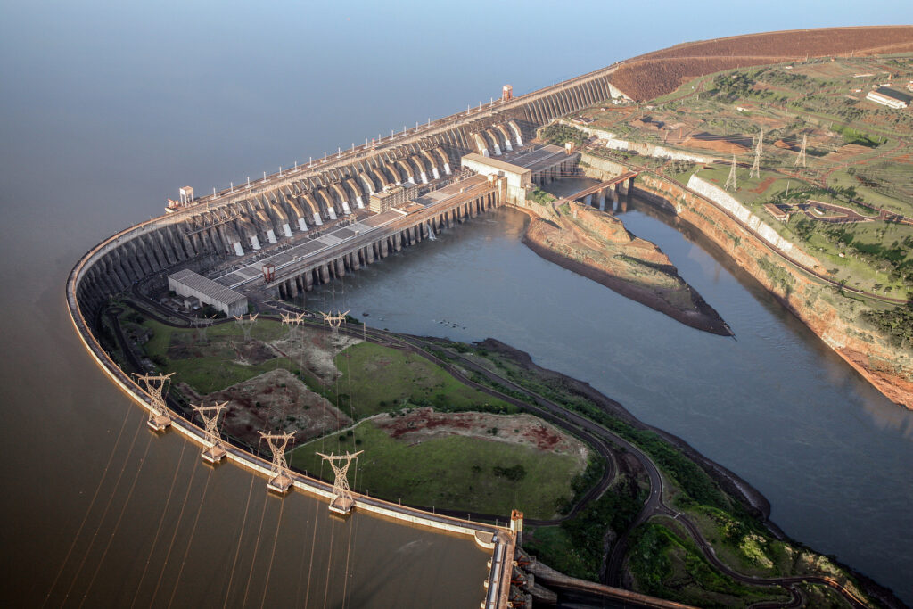 Vista aérea de uma grande represa com água sendo liberada por seus vertedouros e estruturas de transmissão ao redor