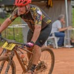 Um ciclista com capacete vermelho pedala em terreno acidentado e enlameado durante prova de mountain bike