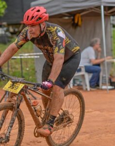 Um ciclista com capacete vermelho pedala em terreno acidentado e enlameado durante prova de mountain bike