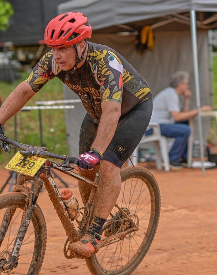 Um ciclista com capacete vermelho pedala em terreno acidentado e enlameado durante prova de mountain bike