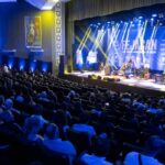 Um grupo de músicos se apresenta em palco diante de plateia em teatro com iluminação azul e amarela