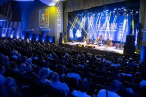 Um grupo de músicos se apresenta em palco diante de plateia em teatro com iluminação azul e amarela