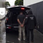 Um adolescente é conduzido para dentro do porta-malas de uma viatura da Polícia Civil em dia chuvoso
