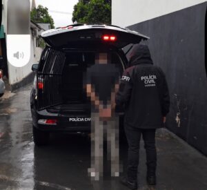 Um adolescente é conduzido para dentro do porta-malas de uma viatura da Polícia Civil em dia chuvoso