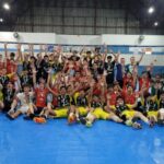 Jovens atletas comemoram em quadra de vôlei, segurando medalhas e levantando as mãos