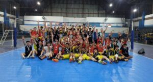 Jovens atletas comemoram em quadra de vôlei, segurando medalhas e levantando as mãos
