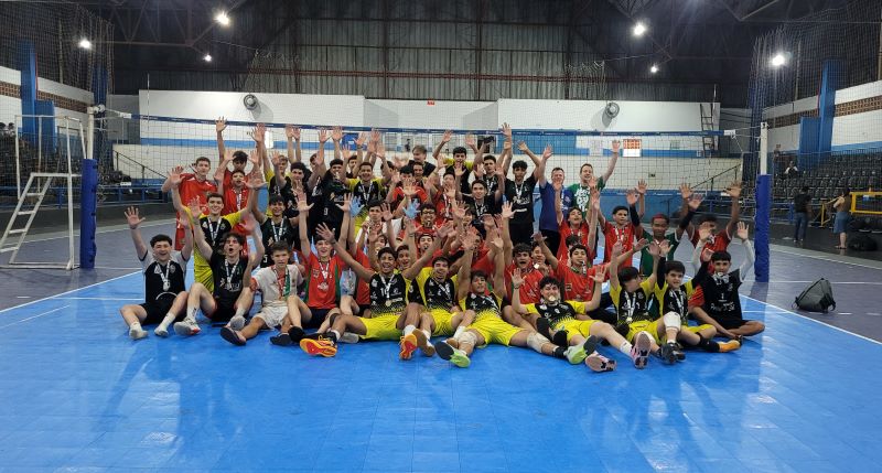 Jovens atletas comemoram em quadra de vôlei, segurando medalhas e levantando as mãos