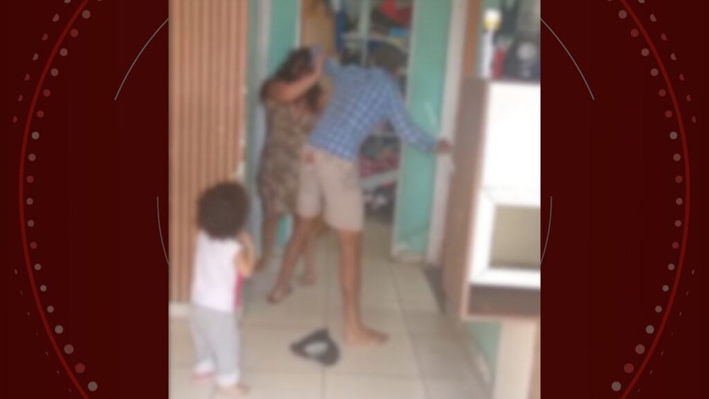 Um homem segura uma mulher pelo pescoço em ambiente interno enquanto uma criança observa ao fundo