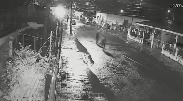 Duas pessoas correm por rua molhada em cena noturna captada por câmera de segurança