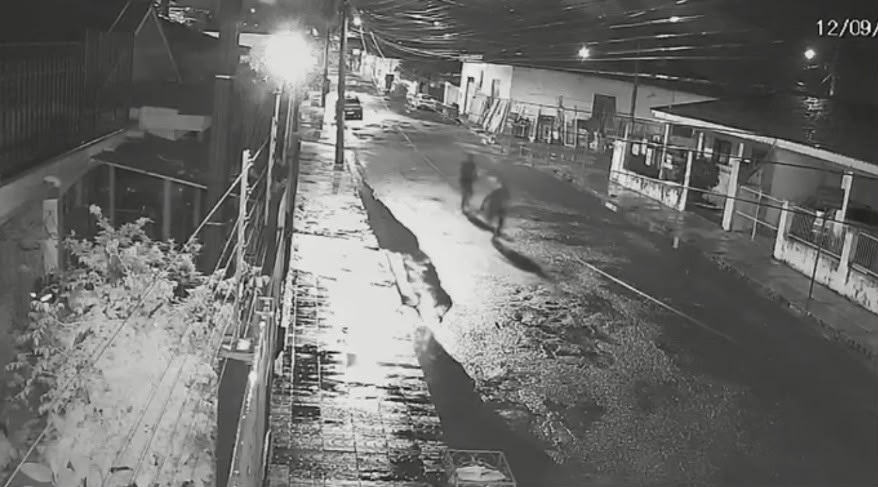 Duas pessoas correm por rua molhada em cena noturna captada por câmera de segurança