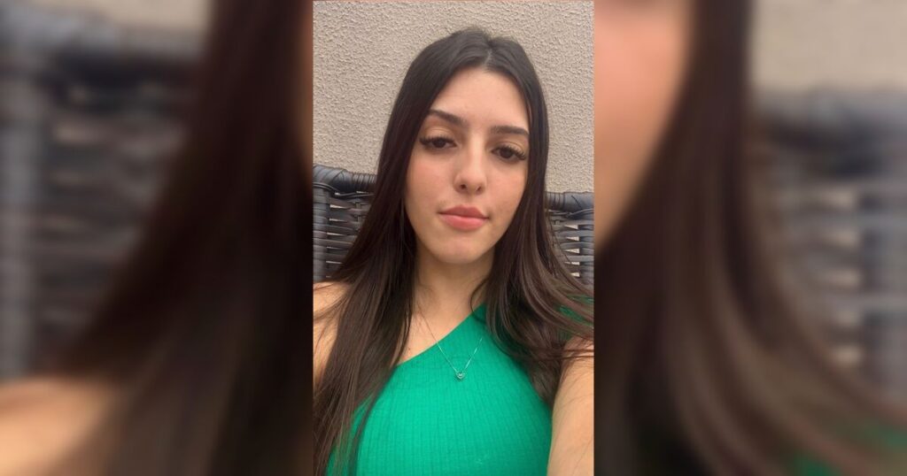 Uma jovem com cabelos longos e camiseta verde posa para selfie em frente a parede clara