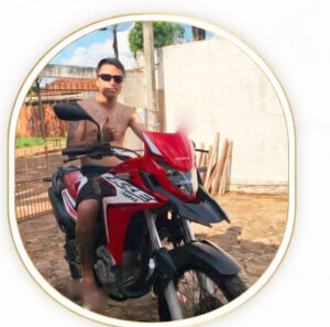 Um homem sem camisa e com óculos escuros sentado em uma moto vermelha em frente a um muro