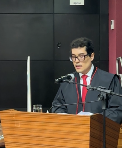 Um homem de toga e gravata vermelha fala em púlpito com microfone em ambiente formal