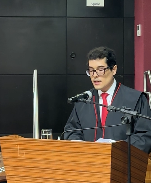 Um homem de toga e gravata vermelha fala em púlpito com microfone em ambiente formal
