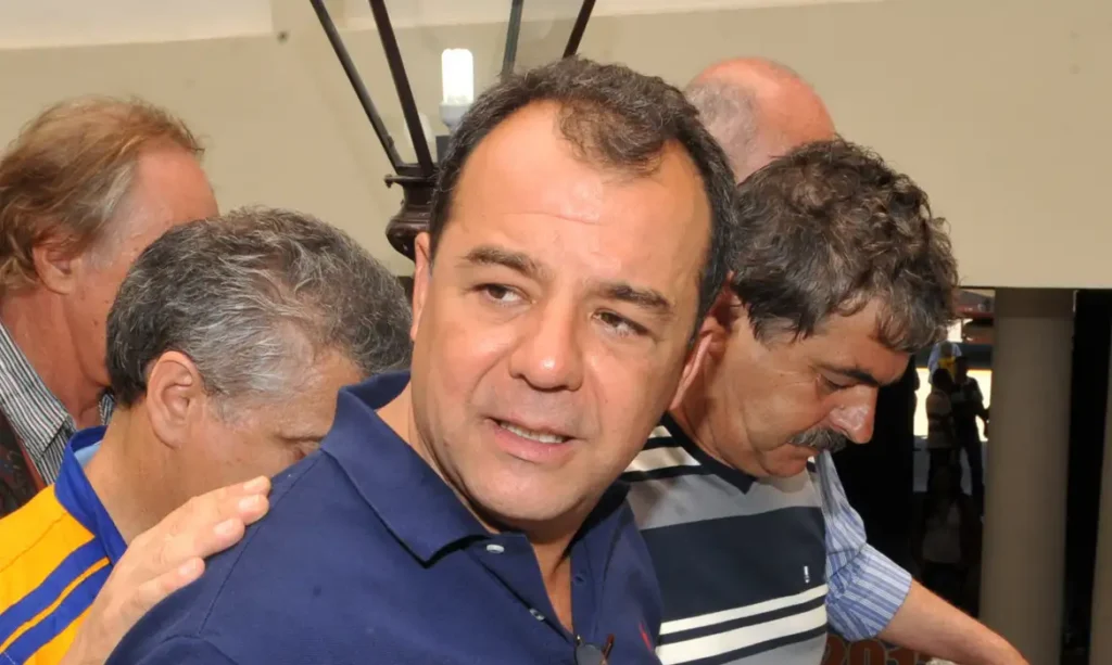 Um homem de camisa azul está cercado por outras pessoas em ambiente interno