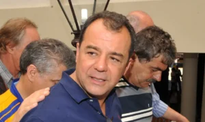 Um homem de camisa azul está cercado por outras pessoas em ambiente interno