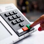 Uma mão pressiona o botão verde em uma urna eleitoral da Justiça Eleitoral