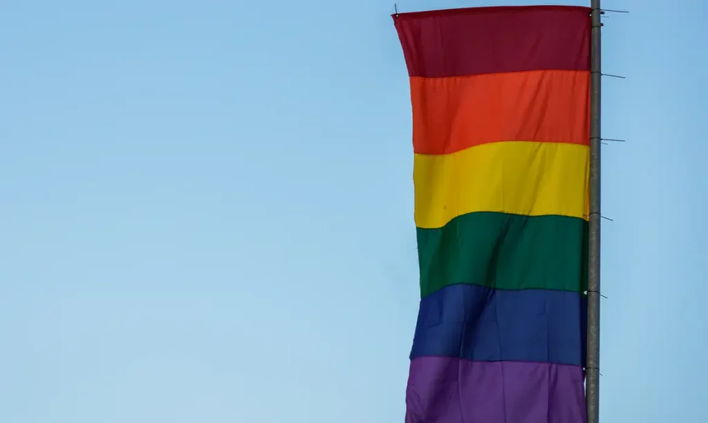 Bandeira com faixas coloridas em tons de vermelho, laranja, amarelo, verde, azul e roxo hasteada em poste contra céu claro