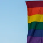 Bandeira com faixas coloridas em tons de vermelho, laranja, amarelo, verde, azul e roxo hasteada em poste contra céu claro