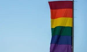 Bandeira com faixas coloridas em tons de vermelho, laranja, amarelo, verde, azul e roxo hasteada em poste contra céu claro