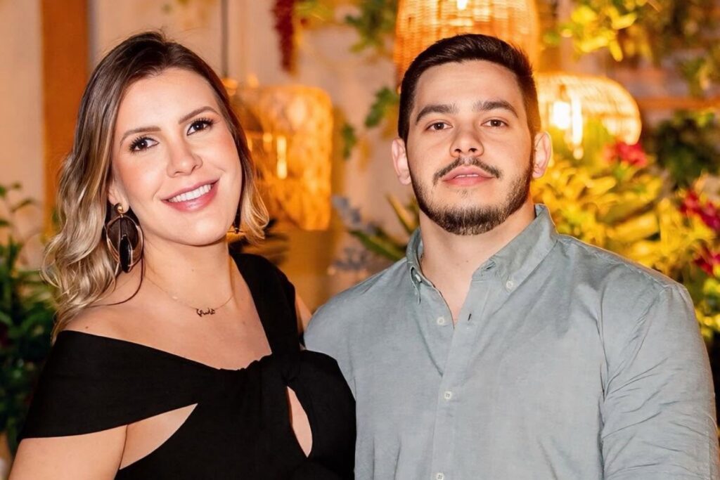 Uma mulher sorridente e um homem posam juntos em frente a decoração com plantas e luminárias