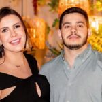 Uma mulher sorridente e um homem posam juntos em frente a decoração com plantas e luminárias