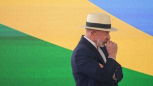 Um homem de terno e chapéu de palha com a mão no queixo diante de painel com cores verde, amarela e azul