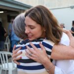 Uma mulher abraça uma idosa em frente a um espaço com cadeiras e pessoas ao fundo