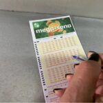 Mão com caneta marca números em volante da Mega-Sena sobre superfície cinza