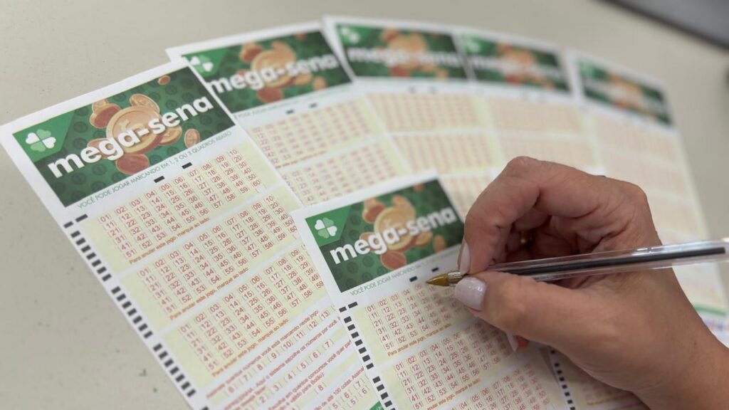 Uma mão segura caneta e marca números em volantes da Mega-Sena sobre uma mesa