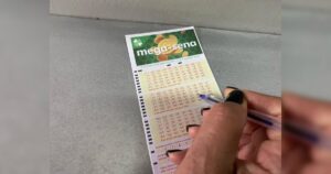 Mão segura caneta e marca números em volante da Mega-Sena sobre superfície cinza