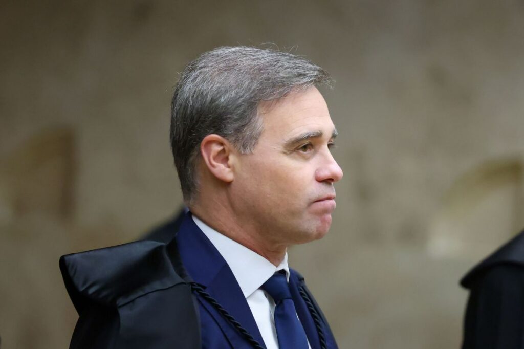 Um homem de terno e capa preta em perfil, com fundo de parede clara