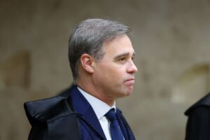 Um homem de terno e capa preta em perfil, com fundo de parede clara