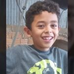 Menino sorridente com camiseta cinza e estampa verde, em frente a parede de tijolos e telha metálica