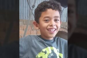 Menino sorridente com camiseta cinza e estampa verde, em frente a parede de tijolos e telha metálica