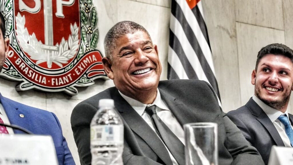 Um homem de terno sorrindo em frente a um brasão e bandeira com listras pretas e brancas