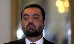 Um homem de terno e gravata com barba olha para frente em ambiente interno com luzes ao fundo
